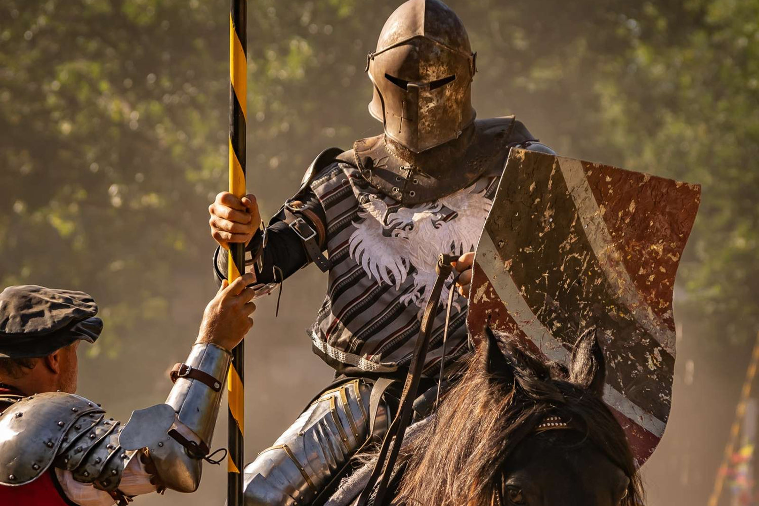Knight on horseback in armor with a banner, engaging with another armored figure outdoors.